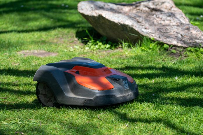 A robotic mower with a red and black casing moves across a grassy lawn, guided by a perimeter wire, with a large rock and some scattered shadows in the background.